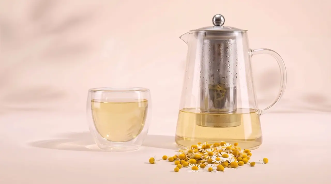 Una taza de vidrio con infusión de manzanilla recién servida, junto a una tetera de vidrio con filtro